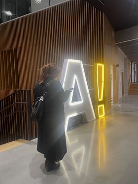 A person in a long black coat in front of a big "A!" light sign.