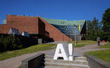 Kuvassa suuri valkoinen Aalto-yliopiston logo on punatiilisen kookkaan rakennuksen edessä portailla.