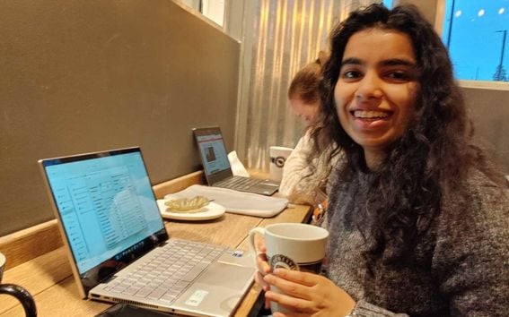 Anubhuti working on a laptop and smiling at the camera holding a big coffee cup.