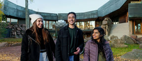 Three students, Amr in the middle, walking on campus with autumn leaves on the ground.