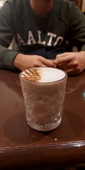 Coffee in a glass on a table in front of a person wearing a shirt with the text "Aalto".